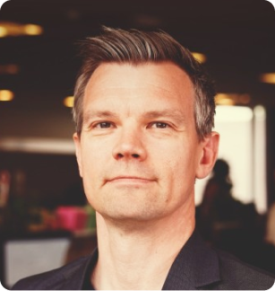 Portrait of a confident man with short grey hair, wearing a dark blazer, set against a blurred office background.