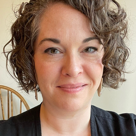Close-up portrait of a woman with curly hair and earrings, smiling softly against a light backdrop, with a wooden chair in view.