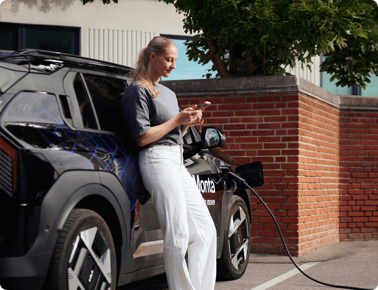 A woman in a grey top and white trousers stands beside a black electric car, looking at her phone while it charges.
